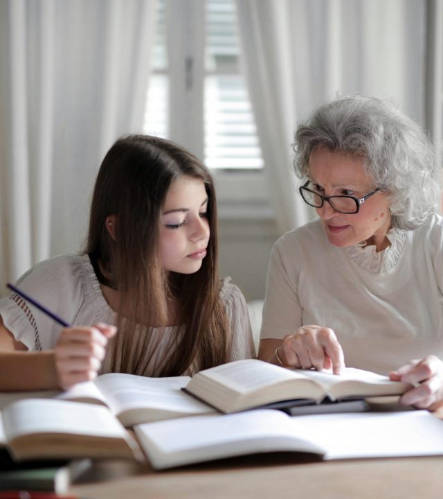 A grandmother helps her teenage granddaughter with homework, surrounded by books indoors.